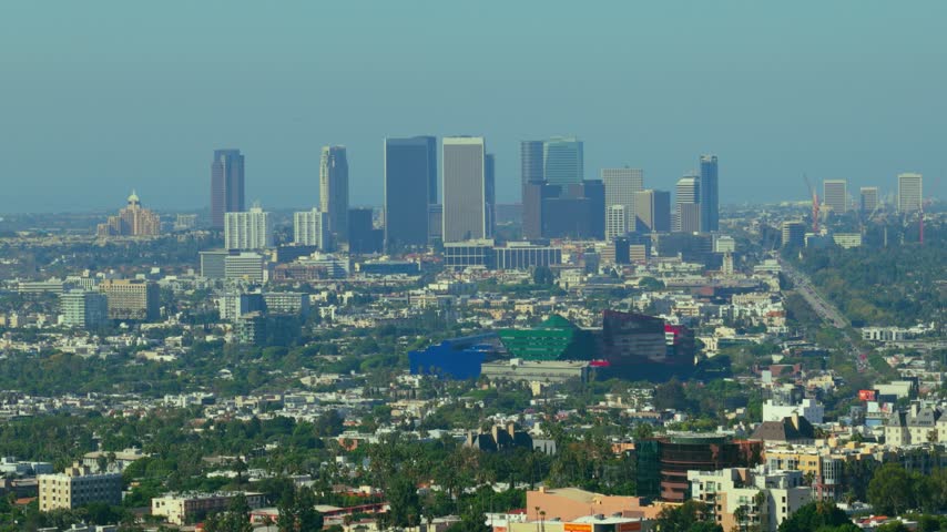 Century City skyscrapers rise above West Hollywood in Los Angeles, California. Telephoto view over palm trees, dense houses and blue, green and red glass buildings on a hazy sunny day. Urban skyline.