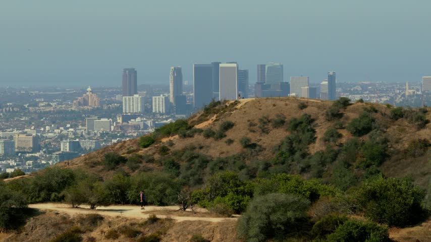 Hikers on a dirt trail across dry hills with the Century City skyline behind. Los Angeles, California on a hazy day. Green shrubs, golden grass and a lone figure on the hilltop. Telephoto view.
