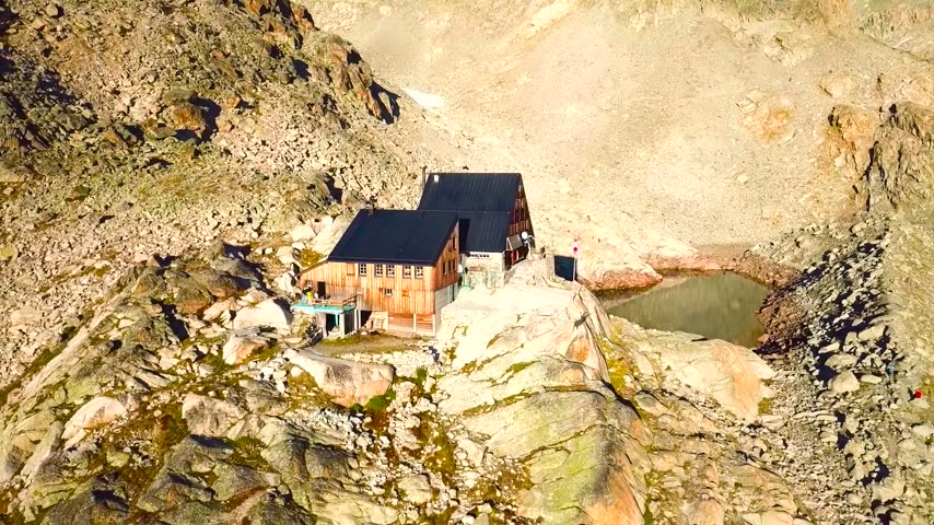 An aerial drone shot of a rustic wooden mountain refuge hut built on a rocky ridge next to a small alpine tarn lake surrounded by rugged crags.
