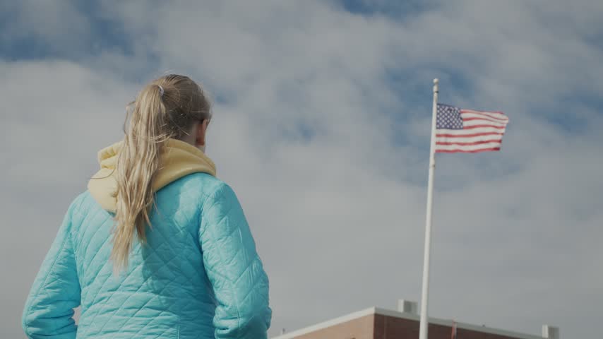 Rear view of school age girl watching United States national flag blowing in wind. Child standing outside brick building during sunny day. Patriotic concept with cinematic motion.