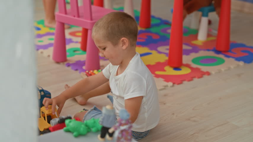 Boy playing with toy truck on foam puzzle mat in indoor playroom, leaning forward and reaching across floor; playful joy.
