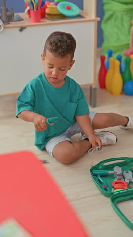Little boy in green shirt sitting on floor holding a plastic screwdriver and toy tool pieces while playing with a green toolset in playroom; curiosity.