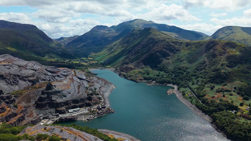 Terraced hillside of an abandoned slate quarry rising above a calm blue lake, steep green valleys and rocky peaks under a bright cloud sky