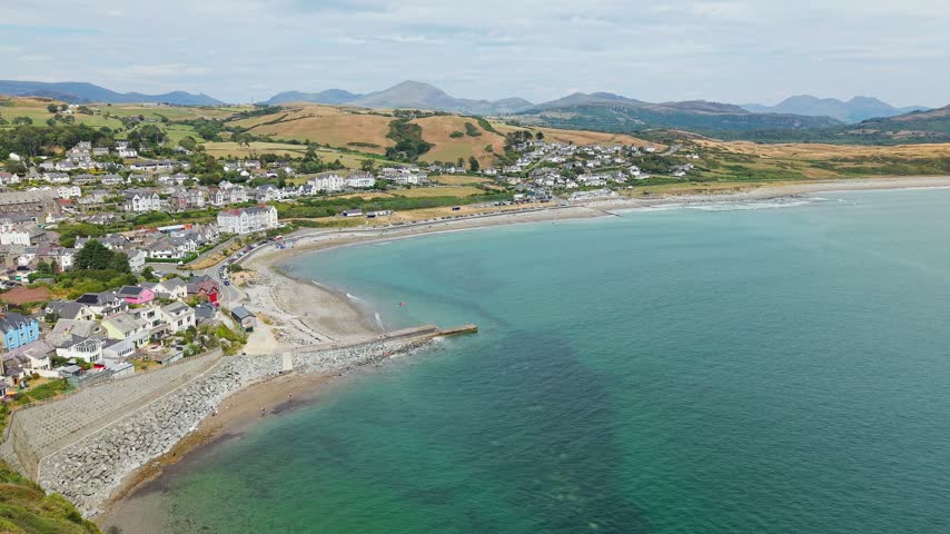 Seaside town and beach along a curved bay, green rolling hills and distant mountain peaks rising behind, aerial view