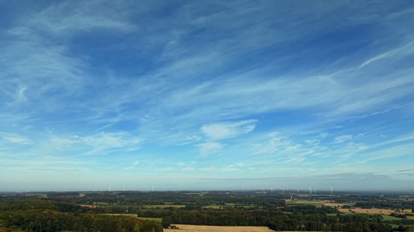 Weather balloon floating in sunny blue sky with wispy cirrus clouds above rural fields, dense forest, distant white wind turbines in America, temperature measurement. Aerial drone wide shot