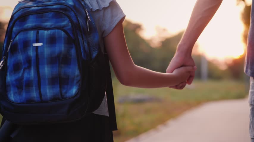 Close up shot captures parent and child holding hands during peaceful walk at sunset. Daughter wearing school backpack explores path together with father cinematic footage.