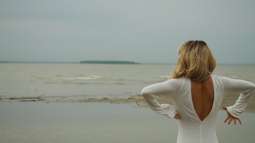 Woman in a white dress raises her arms while standing by the sea. Rear view with open sky and copy space, expressing freedom, relaxation and peaceful coastal lifestyle.