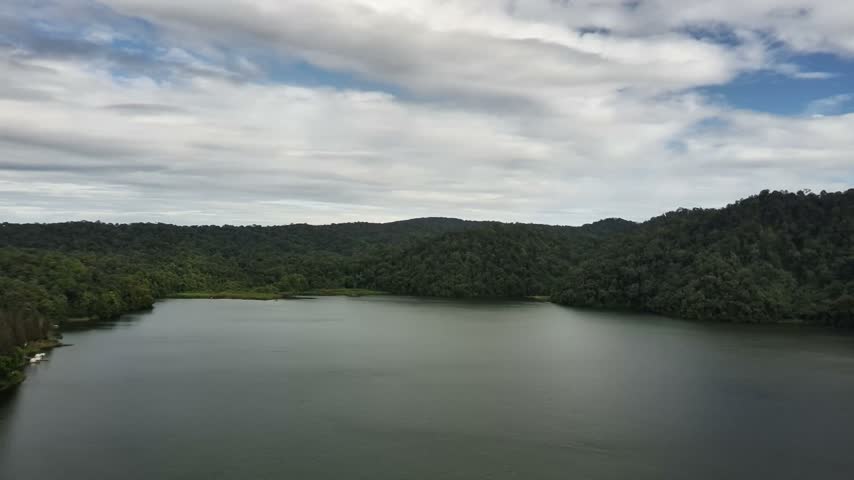 Aerial drone footage rotating counterclockwise above a lakeside valley, revealing calm lake water, lush forests, cultivated land, rural facilities, and surrounding hills beneath a dramatic cloudy sky.
