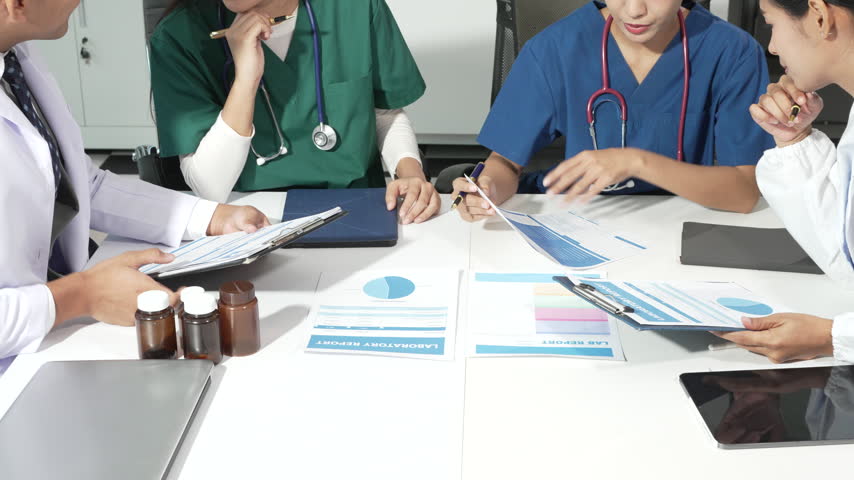 Medical team in scrubs and white coats discussing charts, reports, and medicine bottles at a clinic meeting table.