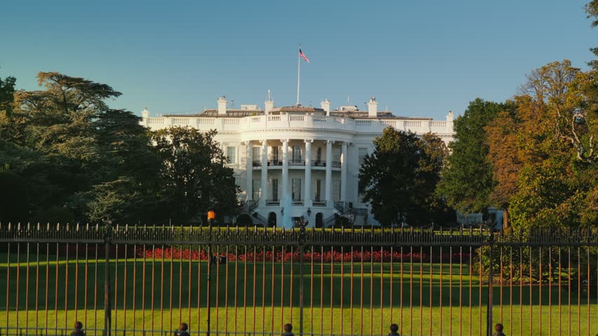 Scenic view of iconic White House architecture featuring lush greenery and prominent US flag waving under clear blue sky during peaceful morning hours in Washington DC.