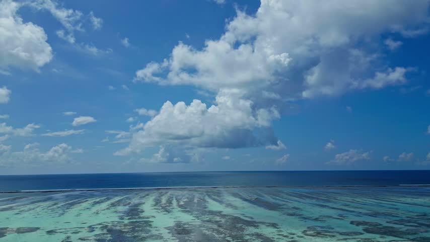 Aerial panorama shows clear turquoise water over coral reef and sandbars, opening to deep blue ocean under a sunny sky with cumulus clouds in tropical Maldives.