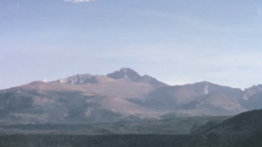 Rocky Mountain National Park features a vast landscape of rolling forested hills and rugged peaks. A slow pan reveals the prominent flat topped summit of Longs Peak under a light blue sky.