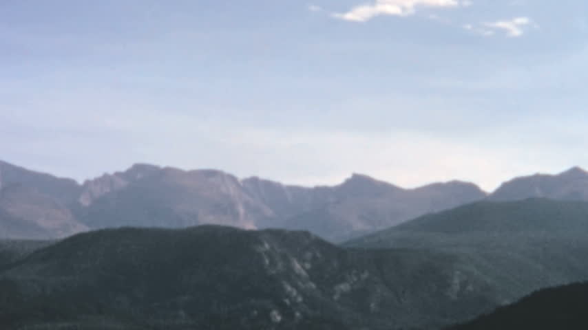 Sweeping Rocky Mountains landscape unfolds during a slow pan across a forested valley in Colorado. Dark rolling foothills rise toward jagged alpine peaks under a blue sky in the 1960s.