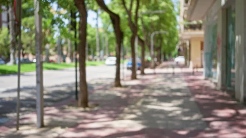 Defocused view of an outdoor street with trees, sidewalks, and a building under a sunny clear sky.