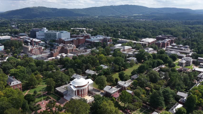 The UVA Rotunda stands amid leafy grounds in Charlottesville, Virginia, with the Lawn behind it and hospital buildings below a wooded ridgeline. Aerial view