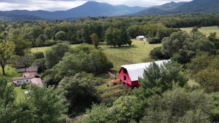 A red barn with a pale roof stands beside pasture in Nellysford, Virginia, surrounded by small outbuildings, mature trees and wooded mountain ridges. Aerial view