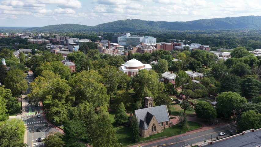 University Chapel and the UVA Rotunda rise among trees in Charlottesville, Virginia, with paths, brick academic buildings and hospital towers beyond. Aerial drone wide shot
