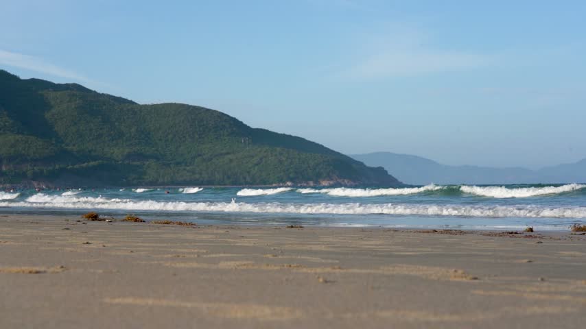 Captivating Shoreline: A Serene Beach Scene Highlighting Gentle Waves, Sandy Coast, and Lush Green Hills Under a Clear Blue Sky