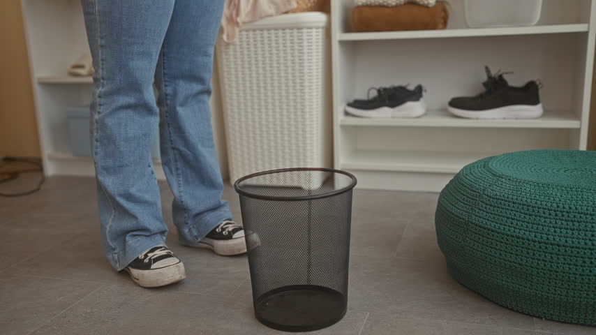 Woman drapes a green shirt over a mesh wastebasket rim while standing in a fitting room building; indecision shopping choice.