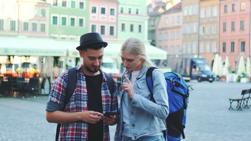 Two young tourists using a smartphone and exploring a vibrant European city square with colorful buildings and outdoor cafes during daytime.