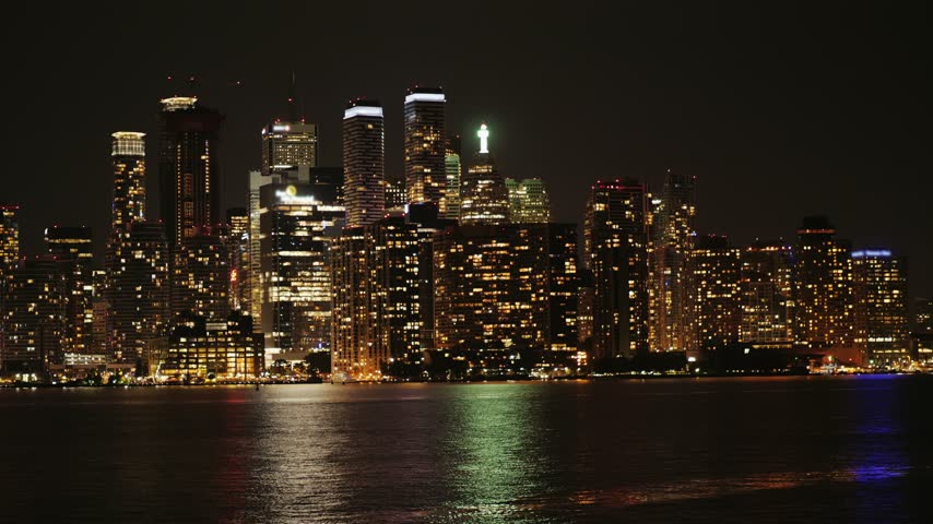 Toronto skyline at night seen from Toronto island across Lake Ontario, capturing illuminated buildings and towers reflecting in water under clear dark sky, cinematic motion footage.