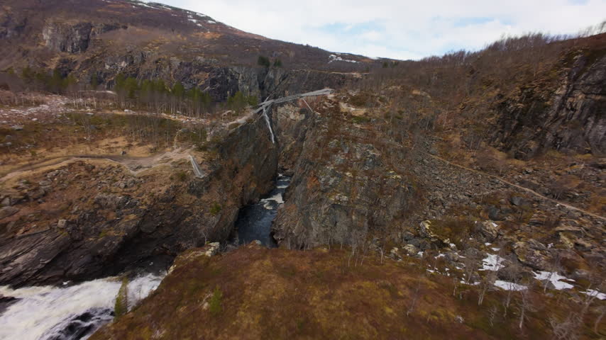 FPV drone flies over the gorge and passes beneath a bridge while approaching Voringfossen waterfall in Norway.