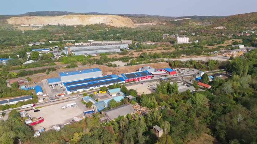 Tatabánya industrial park and factories in Komárom-Esztergom County, Hungary, with warehouses, roads, solar panels and surrounding hills and forest in aerial view.