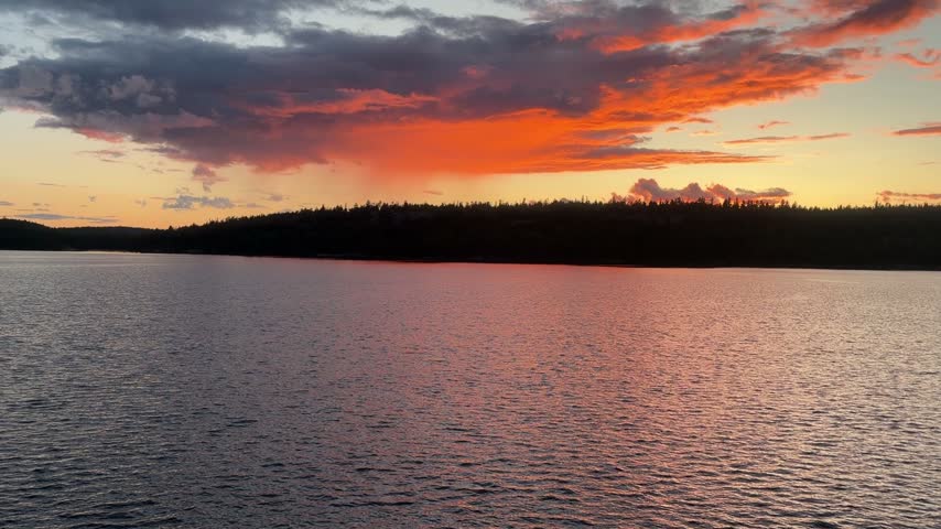Eye level handheld shot looking across calm lake water toward silhouetted forest hills under glowing orange clouds at sunset in Northwest Ontario, Canada.
