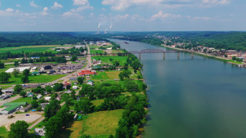 Inland waterway shipping aerial on the Ohio River, barge tow and towboat passing under a truss bridge, railroad trestle and forested banks, West Virginia, USA