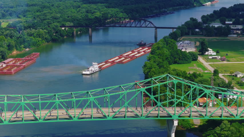 Barge tow Ohio River aerial view, towboat pushing cargo barges under a green truss bridge, railroad trestle and forested banks, West Virginia state line, USA