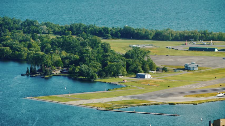 High angle aerial footage showing Billy Bishop Toronto City Airport located on island. Small aircraft is seen taking off from runway surrounded by green trees and blue water of Lake Ontario.