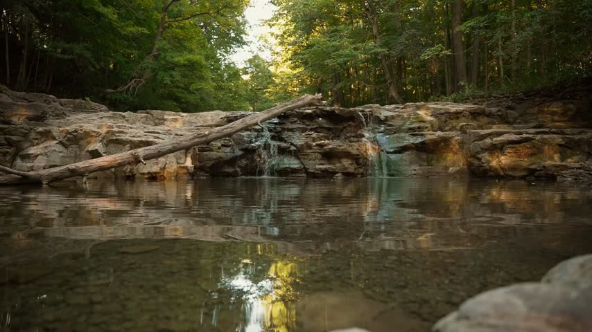 Low-level water surface shot with a picturesque rocky forest waterfall reflected in the foreground stream