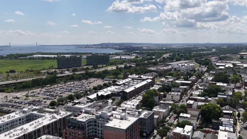 A slow aerial drift over Jersey City, NJ, follows apartment buildings, neighborhood streets and a rail corridor, with green parkland and harbor water extending toward the distant horizon.