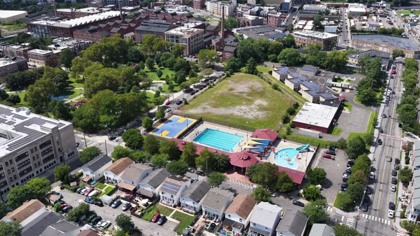 A turning aerial view crosses a park in Jersey City, NJ, revealing green lawns, winding paths and colorful play areas before moving toward nearby residential streets and tightly spaced rooftops.