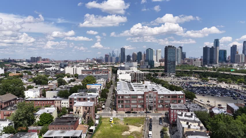 A drone drifts above Jersey City, NJ, revealing a grid of residential streets, apartment buildings, parking areas and modern towers, with the distant Manhattan skyline beneath summer clouds.