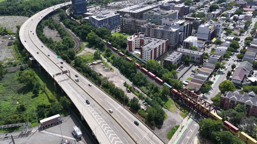 A sweeping drone move follows a curved elevated highway in Jersey City, NJ, revealing rail lines, modern apartments and parking areas before opening toward parkland and distant harbor water.