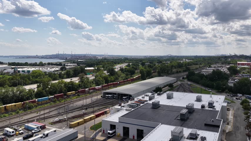 An aerial pan follows rail lines in Jersey City, NJ, passing long industrial buildings, freight cars and service roads, while green trees separate the transport corridor from nearby homes.