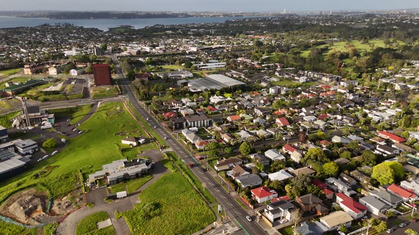 Red brick tower building above residential suburb with houses, main road, industrial buildings, green fields, distant harbour under hazy sky in Mount Albert, Auckland. Aerial drone wide shot