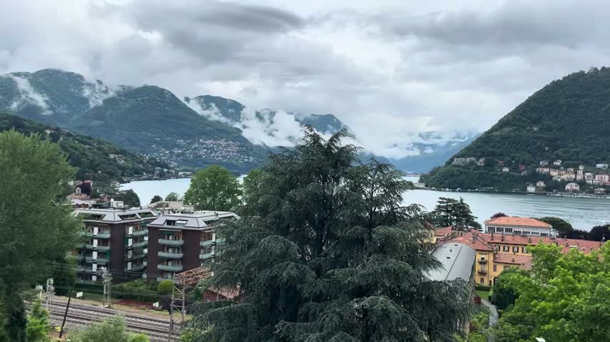 Scenic wide angle view of Lake Como, Italy, with historic town architecture, green hills, and low clouds.