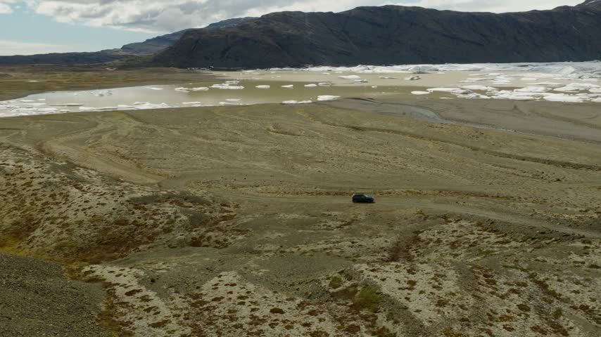 SUV crossing the Heinaberg glacial outwash plain near Heinabergsjokull, Southeast Iceland, with meltwater, glacial ice and dark mountain ridges, drone pullback tracking