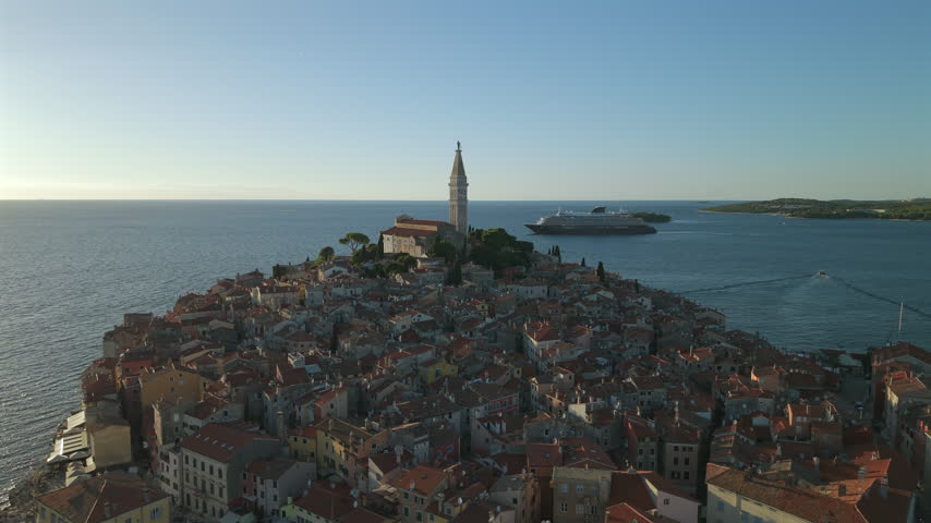 Elevated aerial view over the dense red roofs of Rovinj old town peninsula, Church of Saint Euphemia campanile, and a cruise ship sailing on the blue Adriatic Sea in Croatia