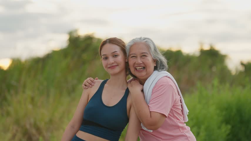 Golden light warms two smiling women embracing in a field. Younger woman wears athletic gear, arms crossed, looking happy. Older woman laughs