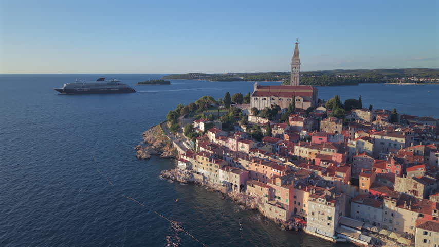 Scenic aerial view of Rovinj old town peninsula featuring the Church of Saint Euphemia bell tower, colorful waterfront houses, and a luxury cruise ship sailing the Adriatic Sea in Croatia