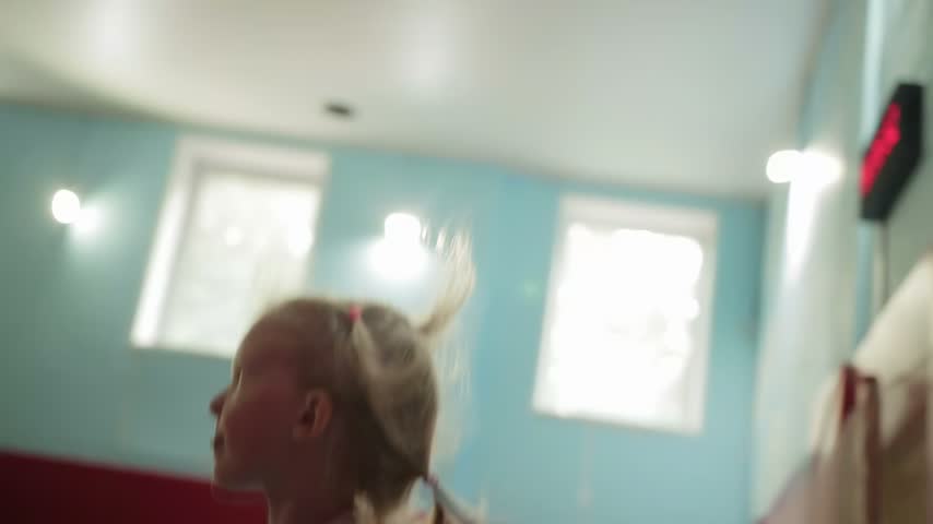 Blonde hair young girl with pigtails actively jumping and having fun in brightly lit indoor play area. Child wears white t-shirt and dark pants. Her movements create motion blur, showing energetic act