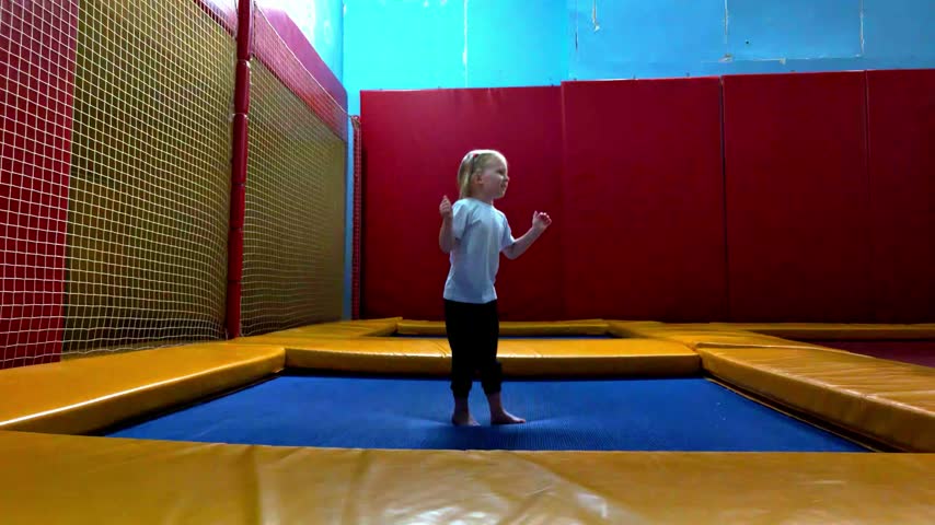 Young blonde girl with pigtails bounces repeatedly on blue trampoline mat, propelling herself high into air. Background shows yellow safety padding and red padded walls of indoor facility.