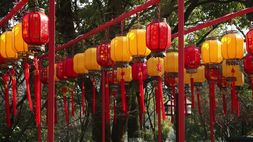 A low angle view captures a series of vibrant red and yellow Chinese lanterns hanging from a red metal frame in an outdoor park setting during the day.