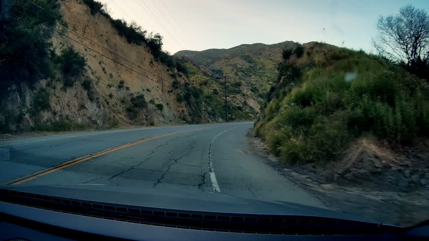 Daytime travel on curved asphalt of Malibu Canyon Road. Rocky terrain flanks road, lush green vegetation, utility poles, power lines visible along winding path. Clear sky above, providing bright light