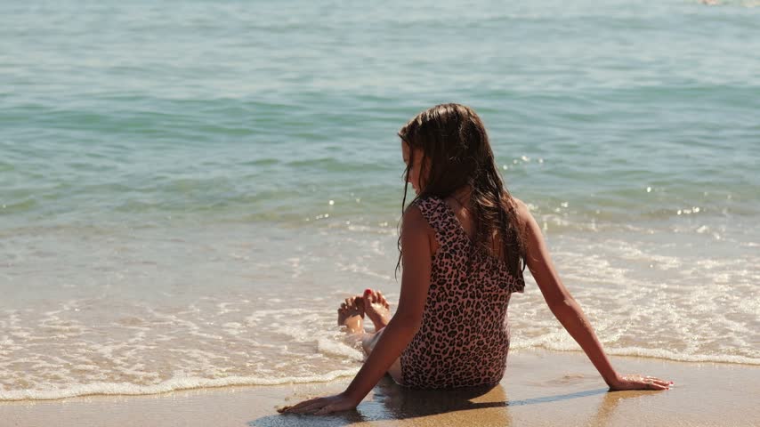 Back view of a young girl in a leopard print swimsuit sitting on the wet shore as gentle sea waves splash around her on a sunny beach day.