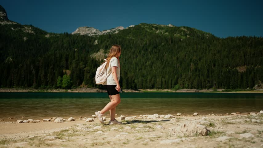 Solo traveler walking along tranquil mountain lake shore, exploring peaceful wilderness landscape with evergreen forest and rocky peaks under clear blue sky