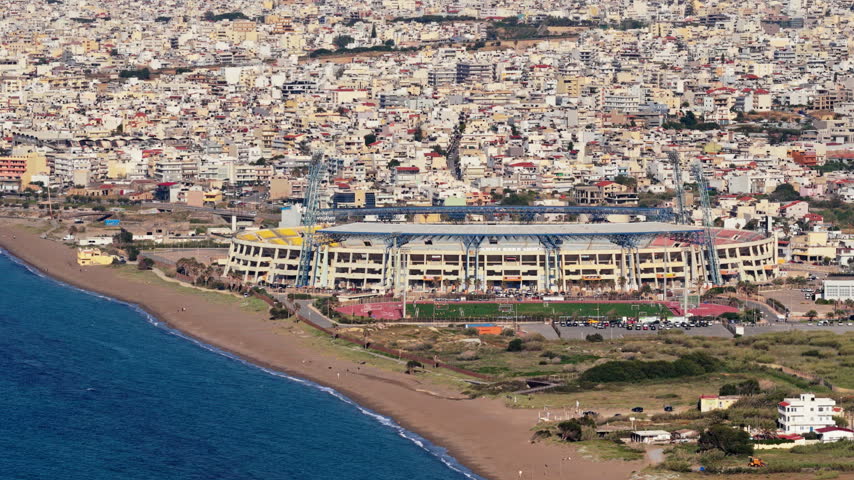 Aerial drone view of the Pancretan Stadium by the sea with the dense city behind it, Heraklion, Greece